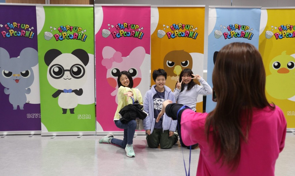 START UP POPCORN!!のカラフルなバナーの前で、子どもたちが笑顔で記念写真に収まっている様子