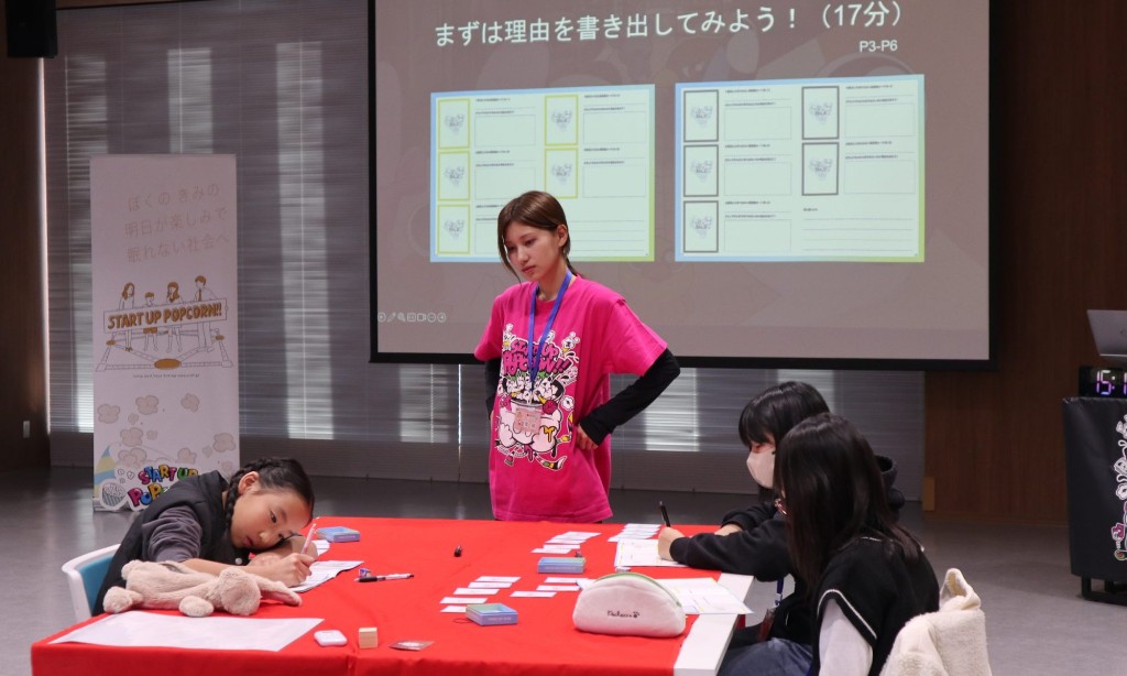 「Work Life Values」のワークシートに、子どもたちが赤いテーブルで記入し、ファシリテーターが見守る様子。スクリーンにはワークの手順が表示されている